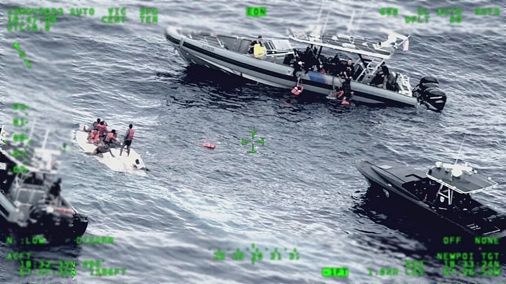 Several people stand on a capsized boat as other passengers are collected by a rescue boat