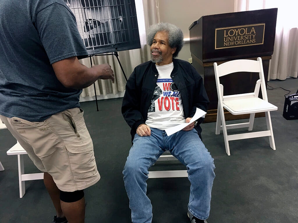 Former prisoner Albert Woodfox sitting in a white chair, wearing a shirt promoting voting