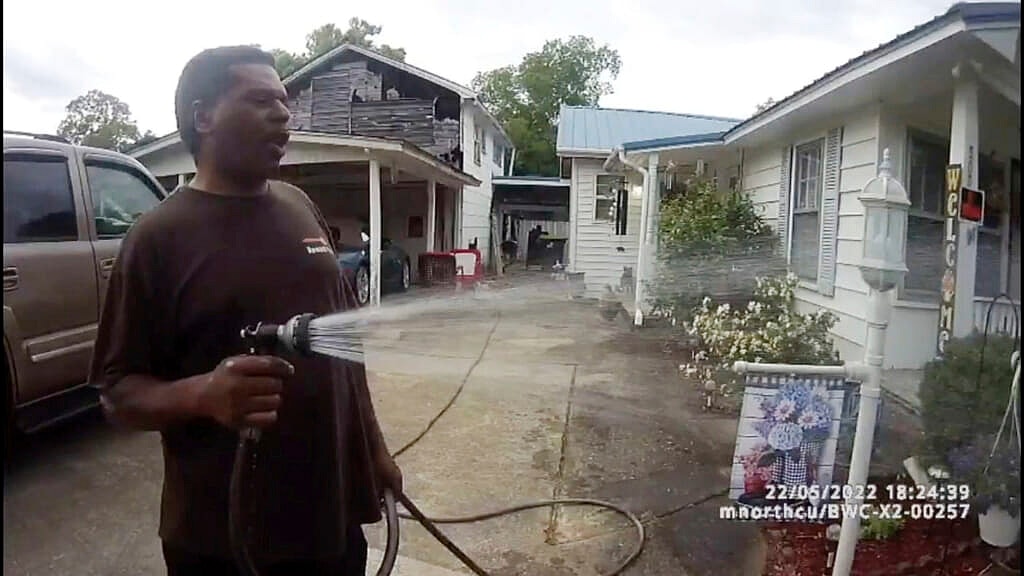 Michael Jennings, in a brown shirt, sprays water from a hose in front of a white house