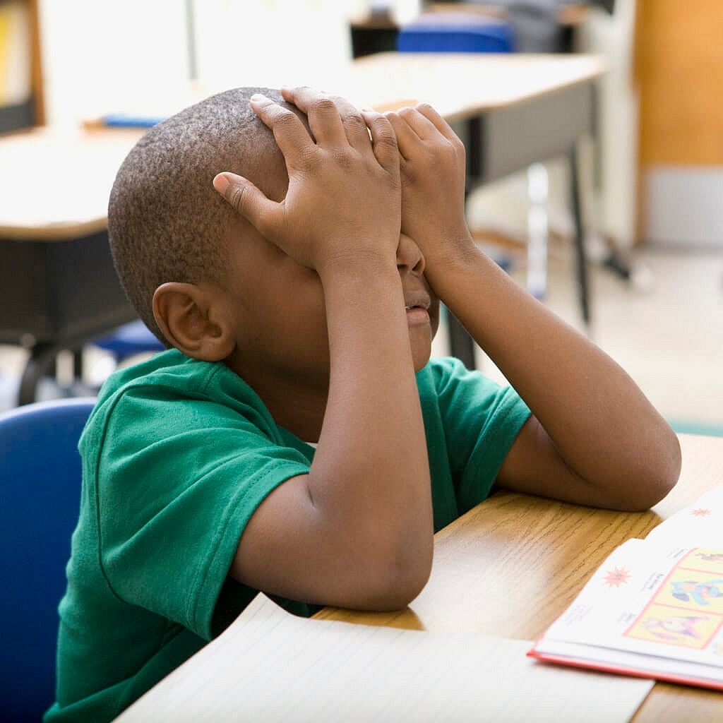 A boy sits at a desk covering his eyes