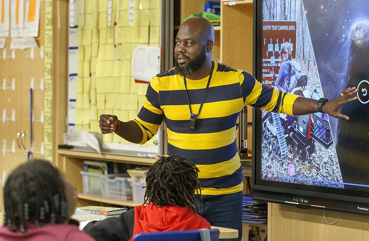 Educator Tyler Wright, wearing a yelllow-and-gray striped top, gestures to a large-screen display