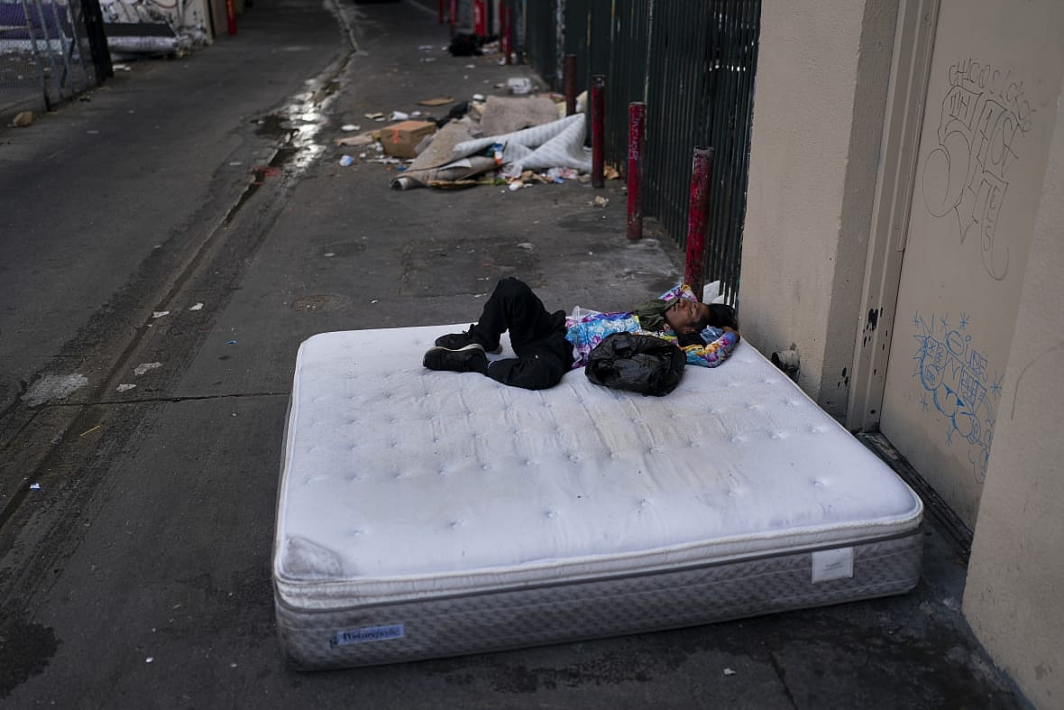 A homeless man wearing a multicolored shirt and black pants sleeps on a discarded mattress