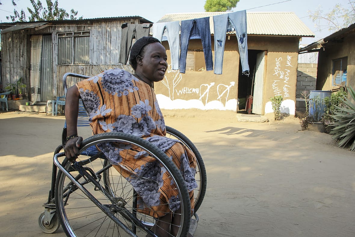 Susan Samson wearing a gold patterned dress uses a wheelchair in front of a roughly made house