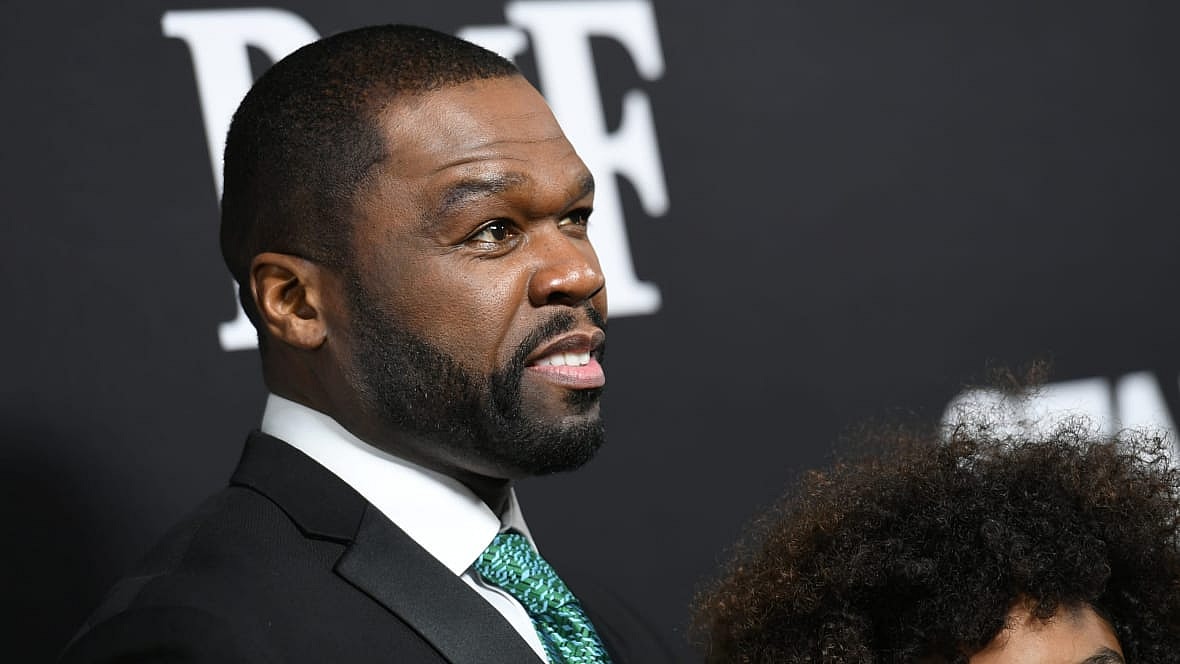 Hip-hop star Curtis “50 Cent” Jackson stands before a black background wearing a black suit and green tie