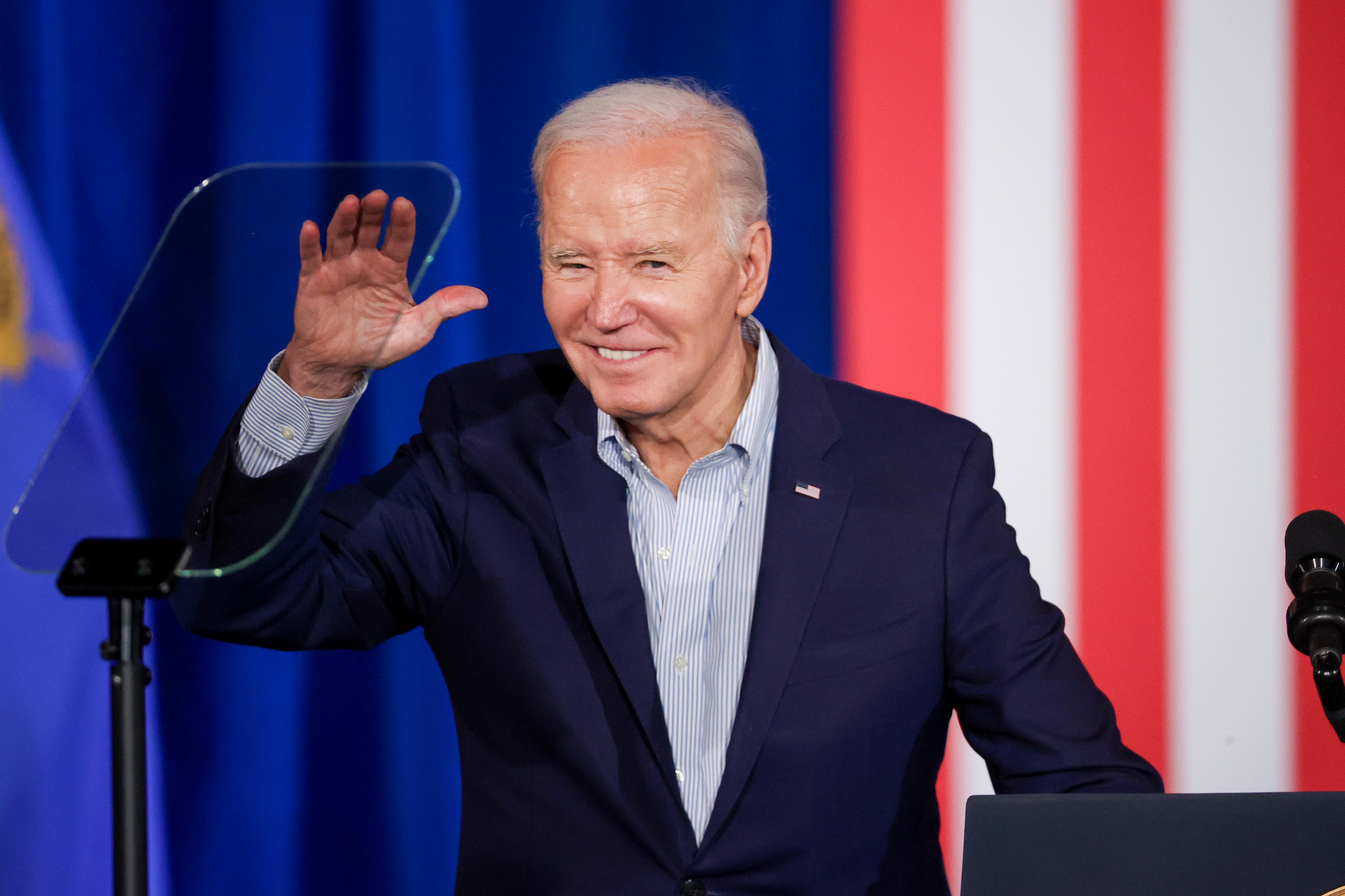 President Joe Biden, wearing a navy jacket and blue-striped shirt, waves from behind a podium