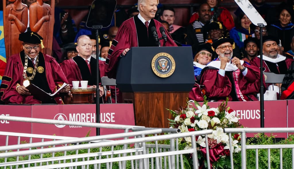 President Joe Biden stands behind a podium wearing a dark red graduate robe over a black suit and red tie