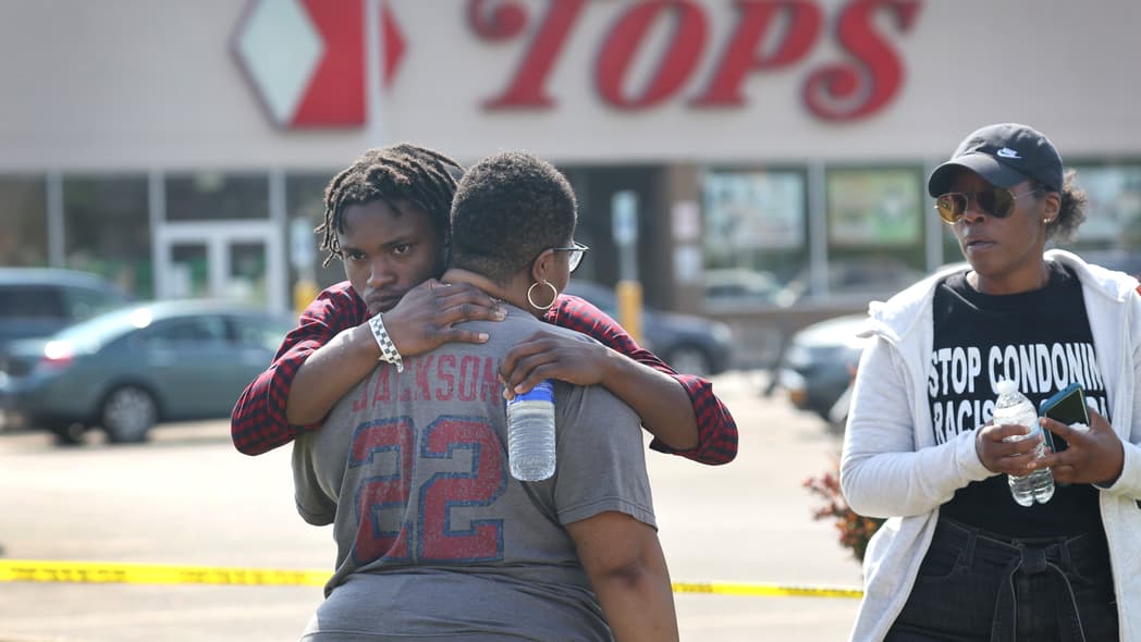 Two people embrace outside Tops market while a person holding a water bottle looks on