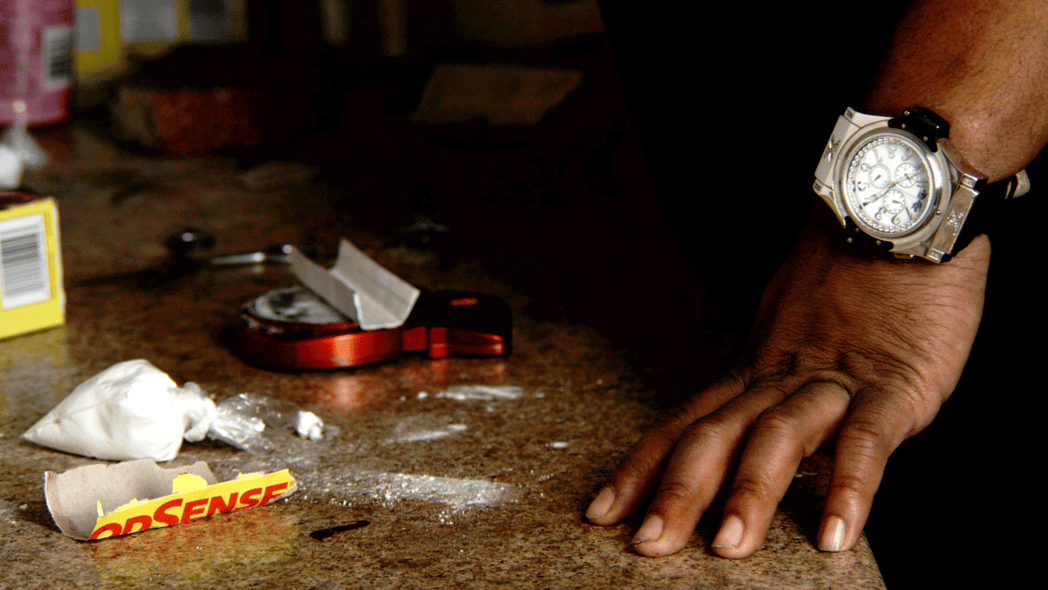 Powder, a scale and other drug paraphernalia are shown on a marble tabletop near a man's hand wearing an expensive watch
