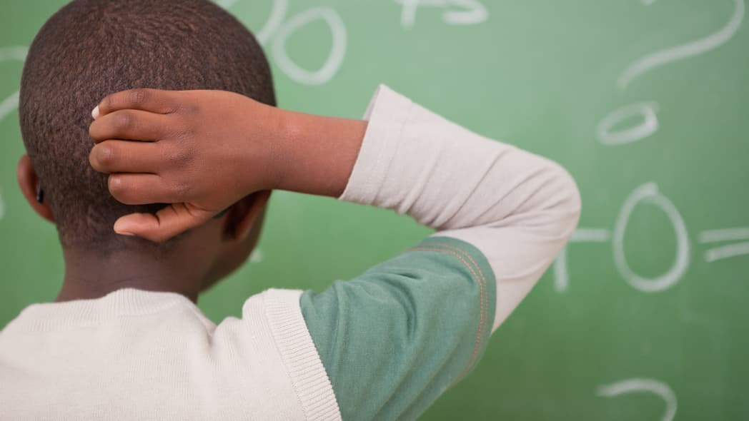 A young boy scratches the back of his head while looking at a green chalkboard
