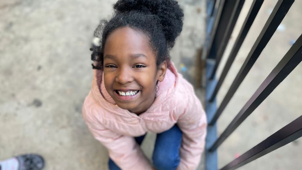 A young girl looks up, smiling, wearing a pink coat and blue jeans