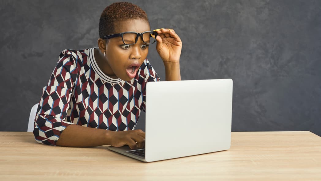A woman lifts her glasses as she looks at a laptop screen in surprise