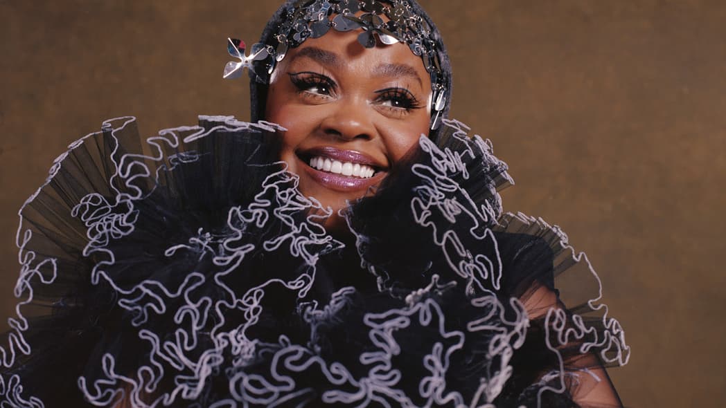 Jill Scott smiles in headshot featuring black and lavender ruffled scarf and hat/headpiece