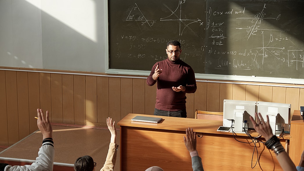 Students raise their hands as a teacher stands behind a desk
