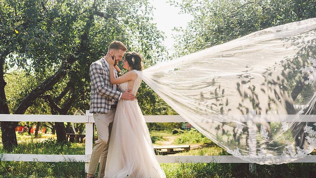 Groom and bride in flowing veil outdoors near a fence