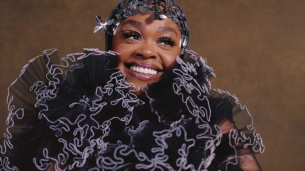 Jill Scott smiles in headshot featuring black and lavender ruffled scarf and hat/headpiece