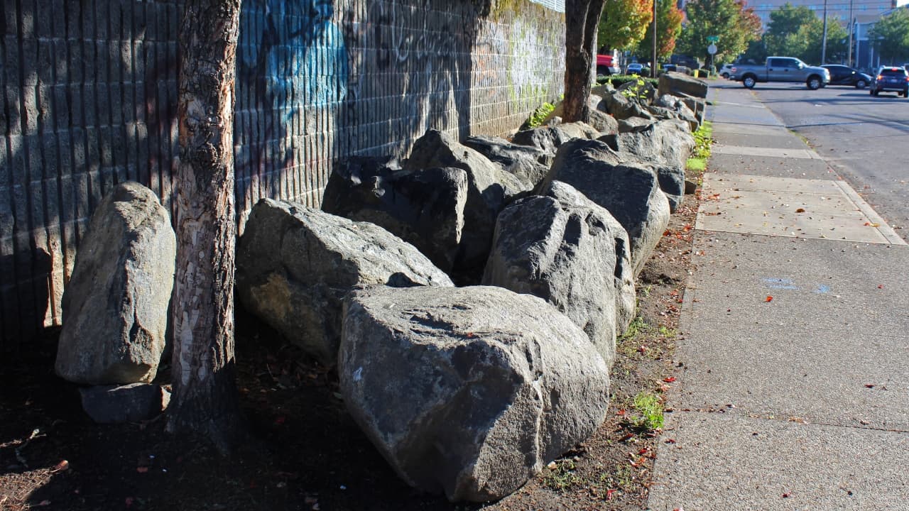 Large boulders on a strip of land between a sidewalk and a parking lot wall