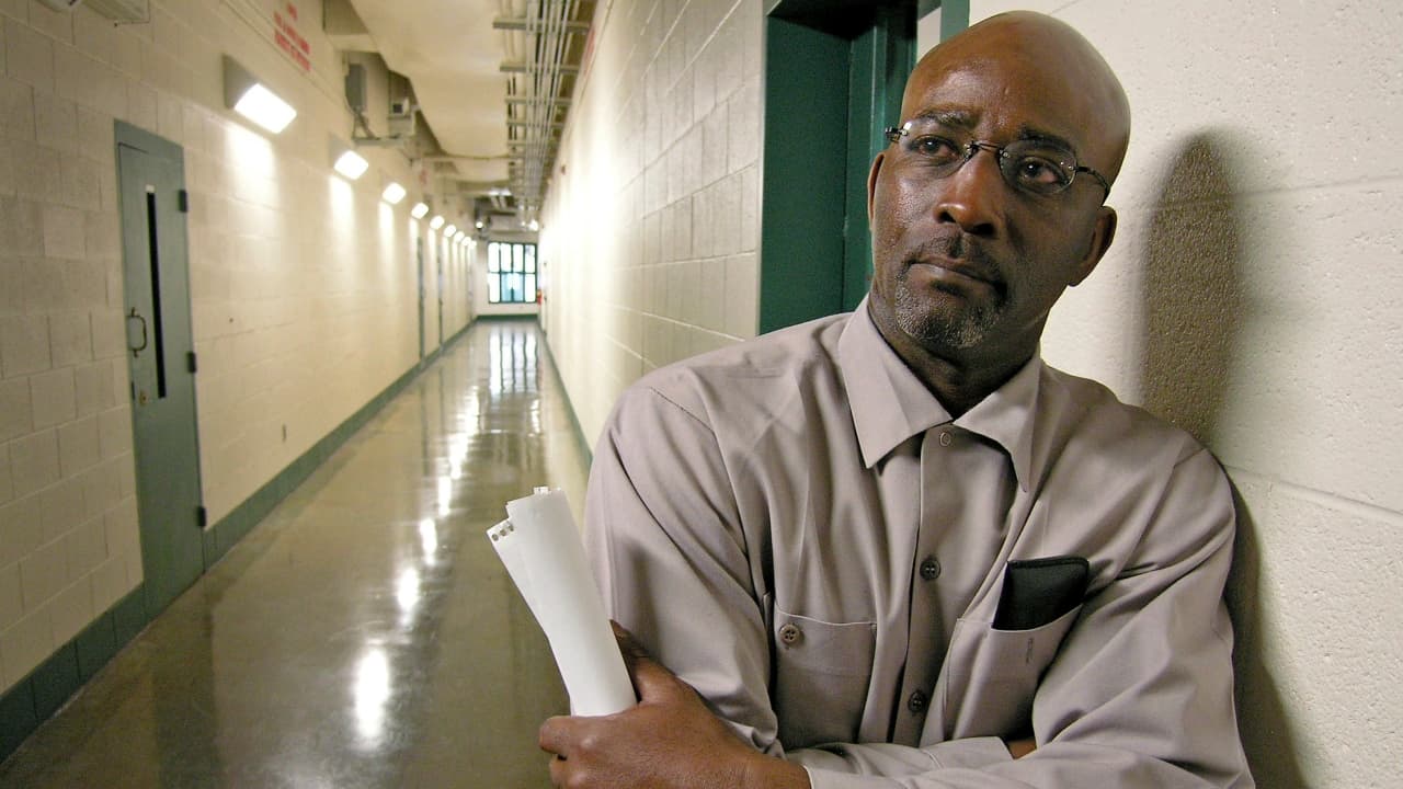 Ronnie Long leans against the wall in a hallway holding a rolled-up document, wearing a light brown shirt