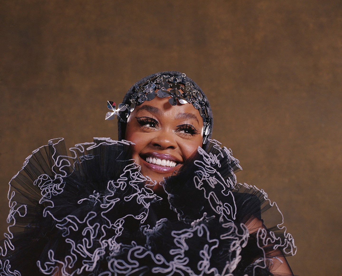 Jill Scott smiles in headshot featuring black and lavender ruffled scarf and hat/headpiece