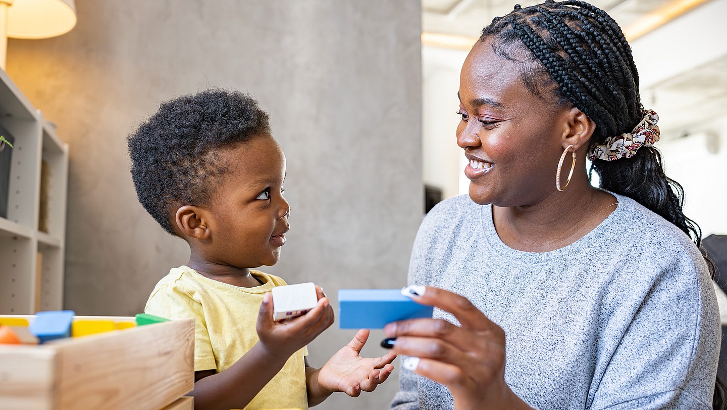 A small child and a woman, each holding a block, smile at each other