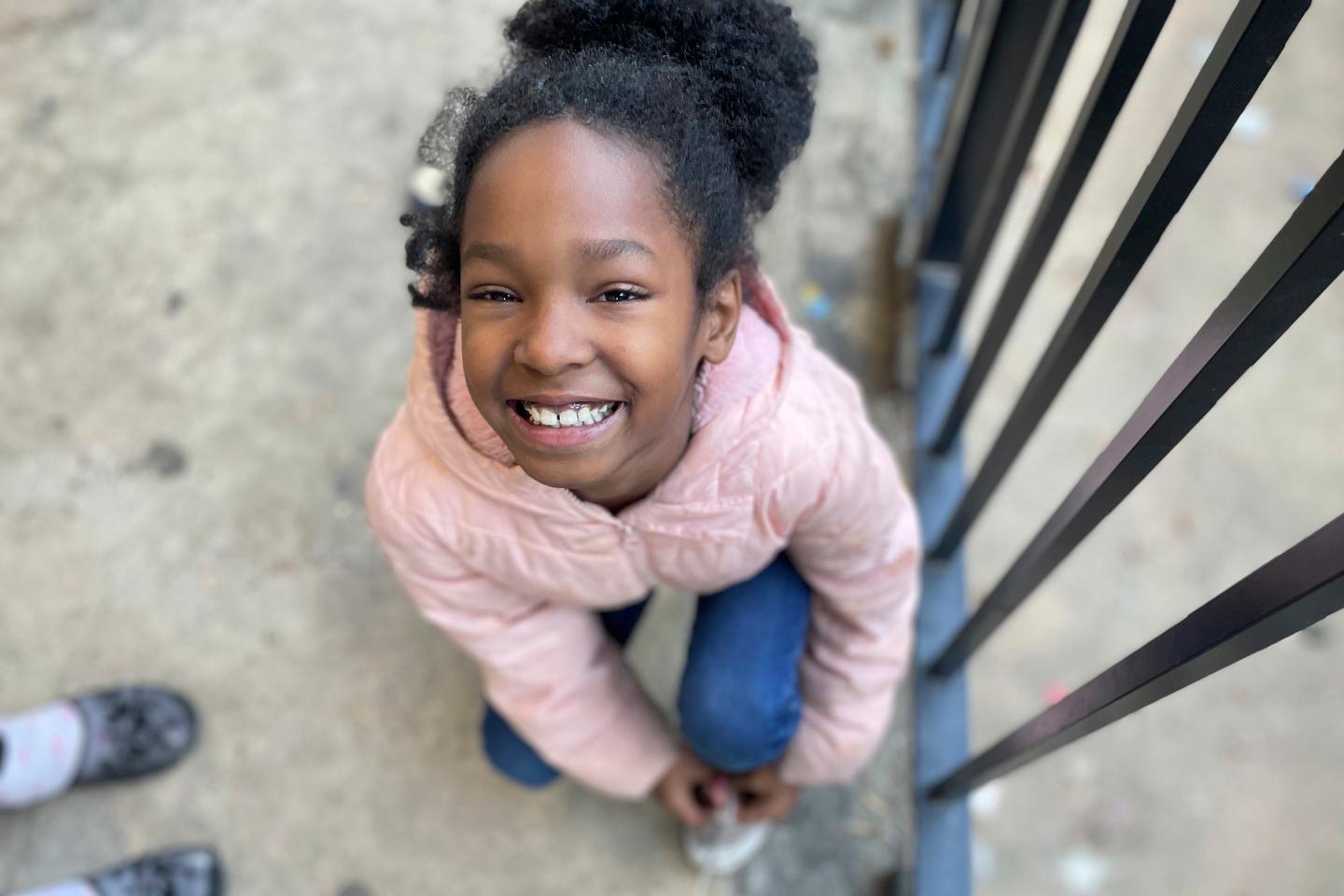 A young girl looks up, smiling, wearing a pink coat and blue jeans