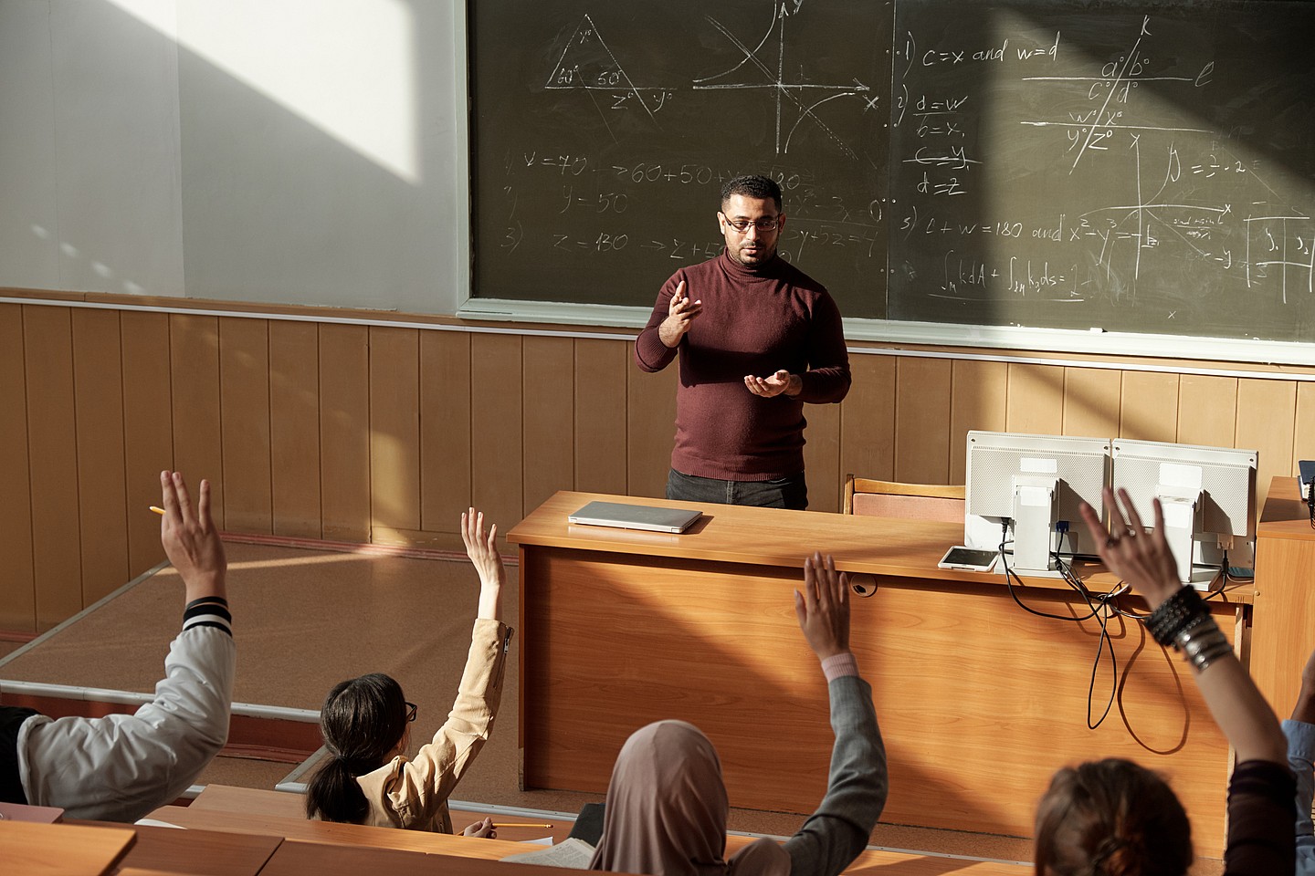 Students raise their hands as a teacher stands behind a desk
