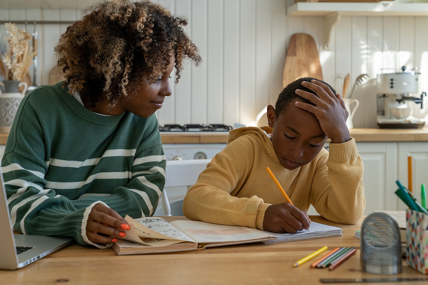 A woman watches a young boy writing in a book