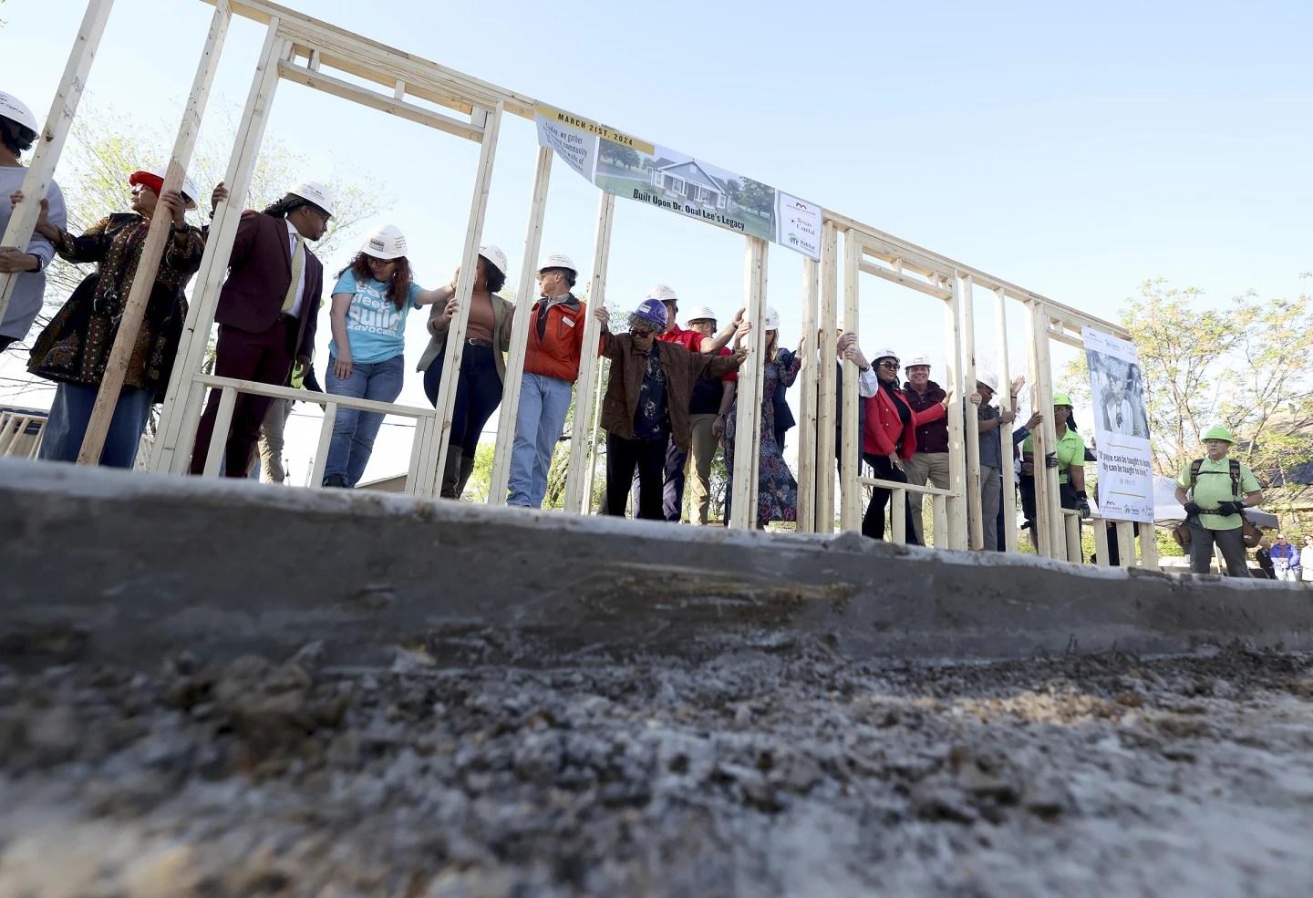 Opal Lee, wearing a purple hard hat and brown coat, stands in the center of the framework of her new home