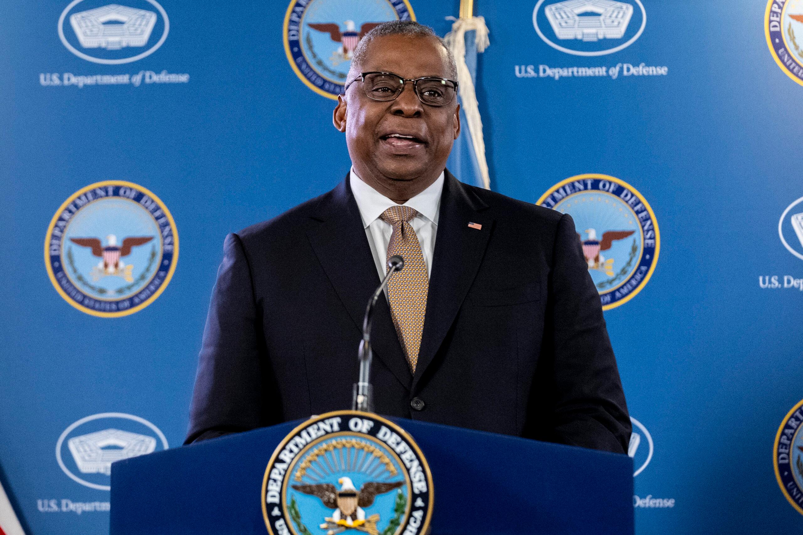 Secretary of Defense Lloyd Austin stands at a podium wearing a navy jacket and brown patterned tie