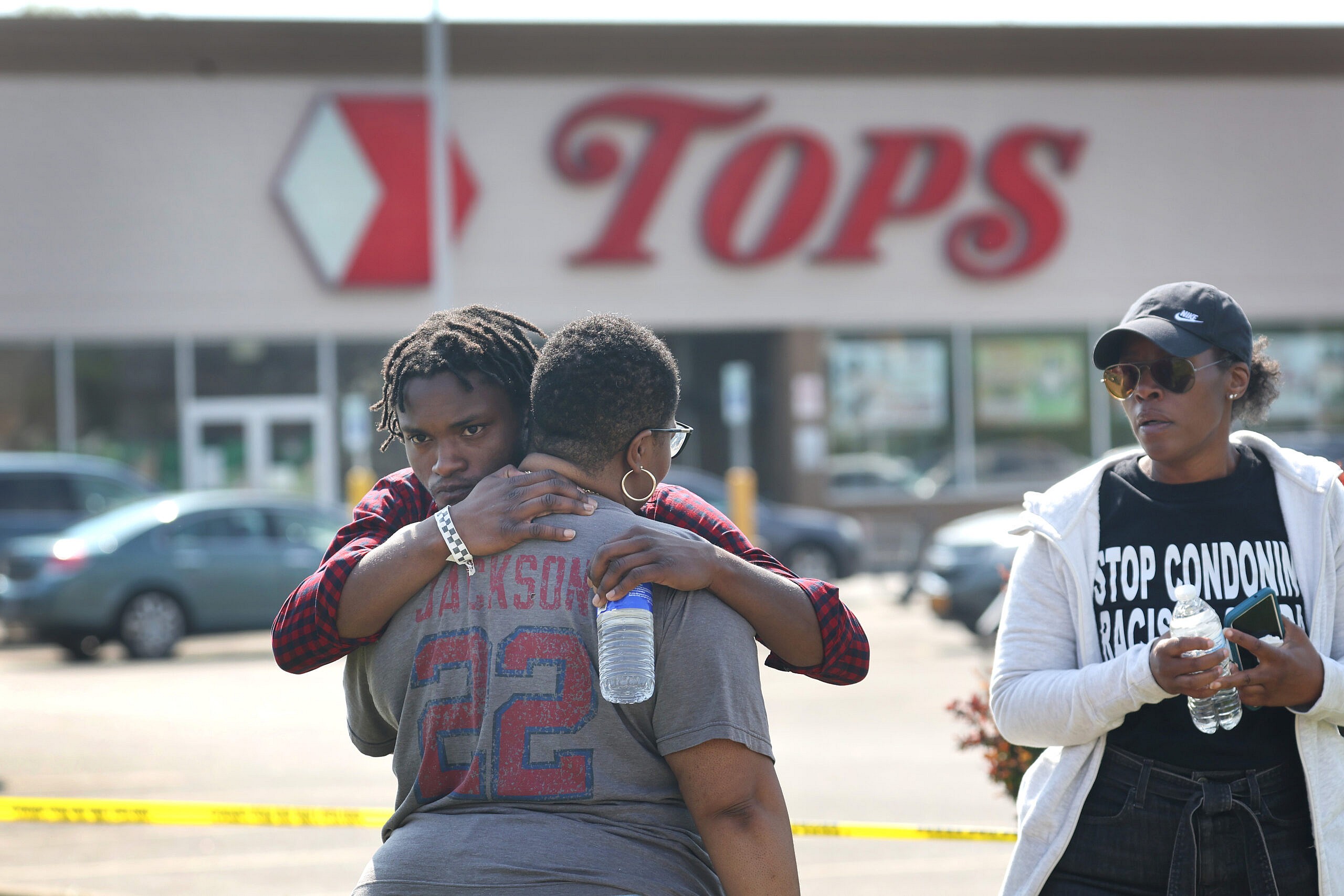Two people embrace outside Tops market while a person holding a water bottle looks on