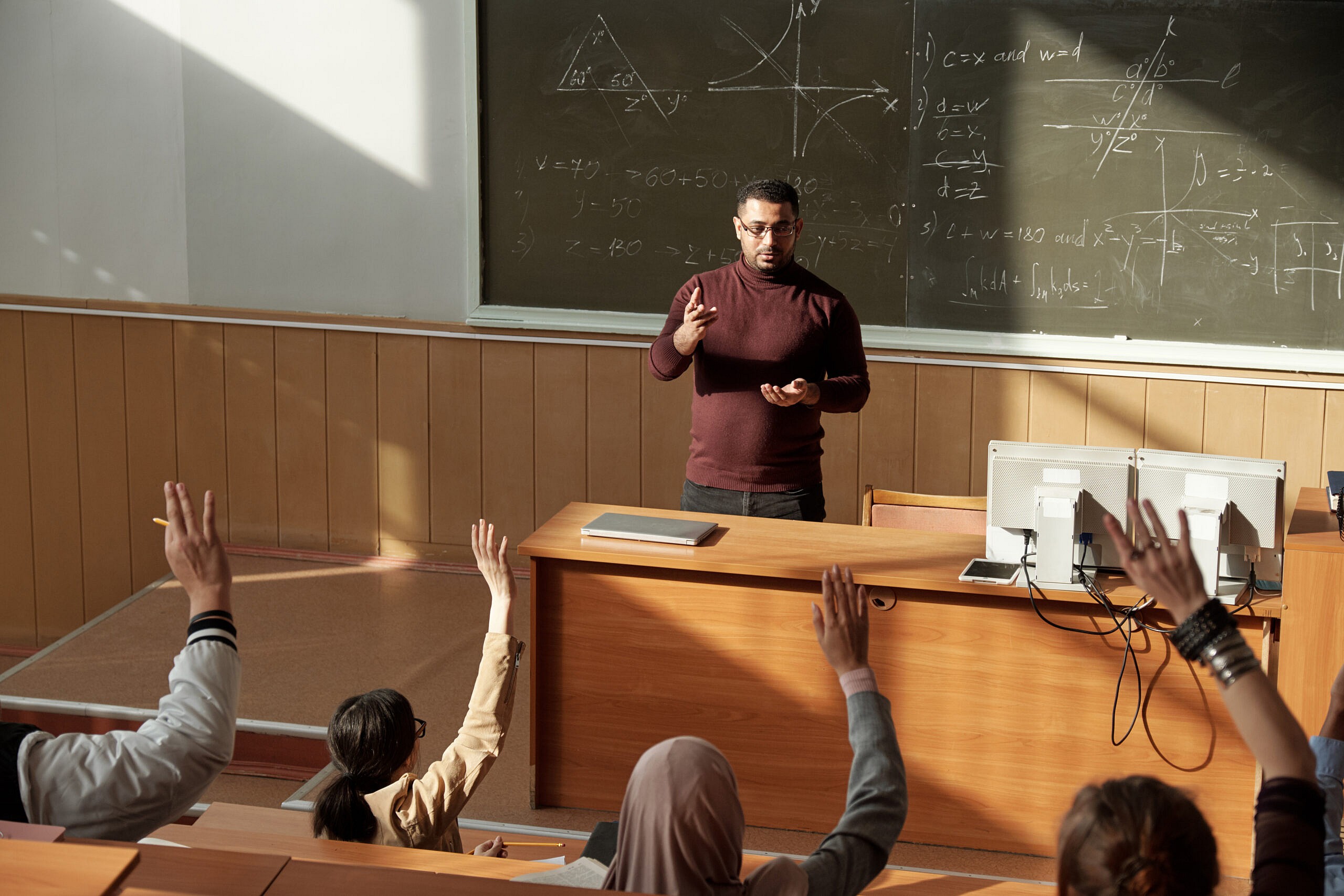 Students raise their hands as a teacher stands behind a desk