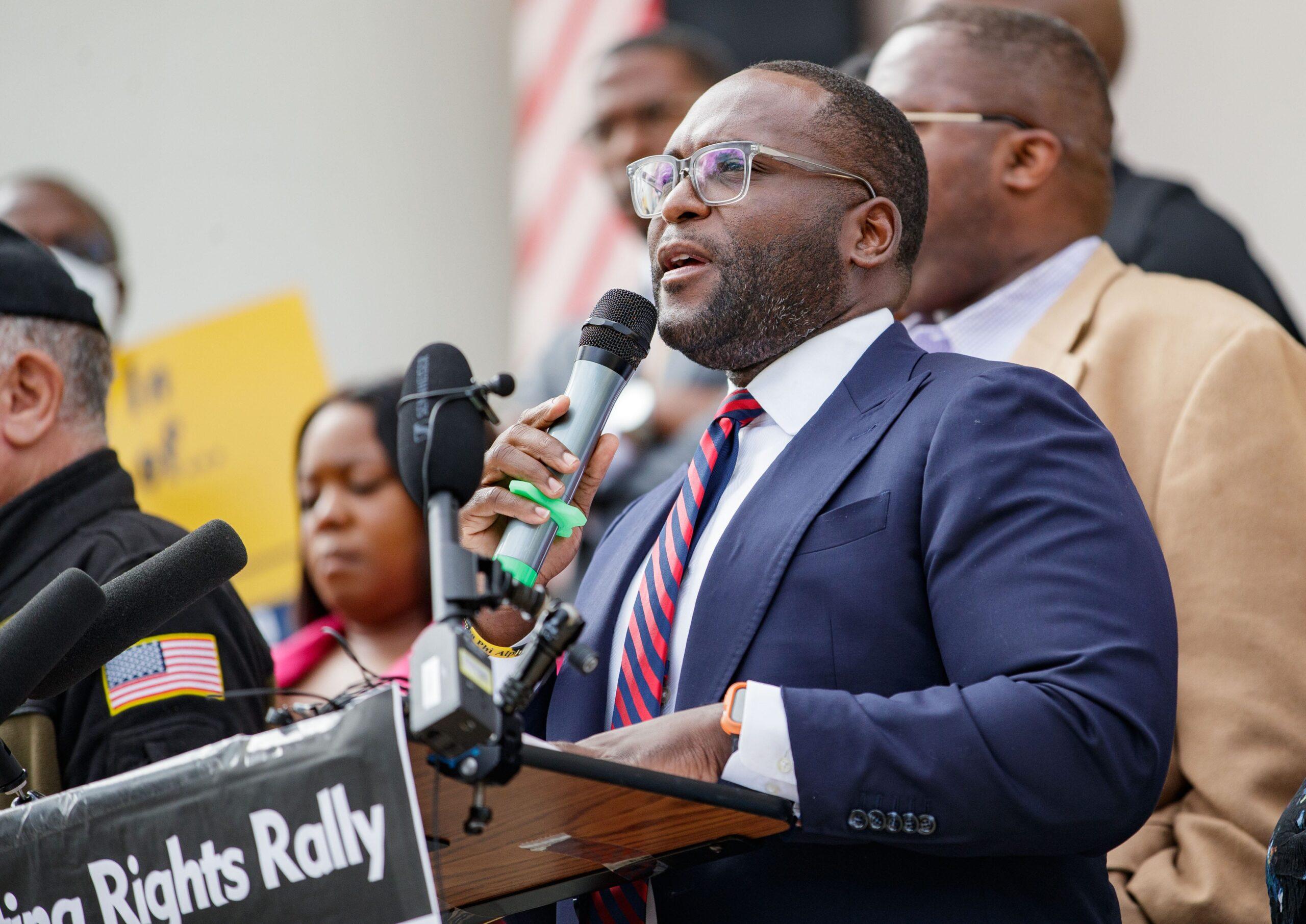 Florida State Senator Shevrin Jones at a podium wearing a blue jacket and red-and-blue striped tie