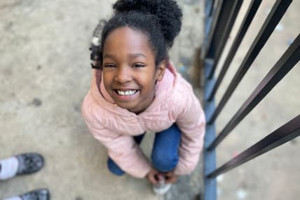 A young girl looks up, smiling, wearing a pink coat and blue jeans