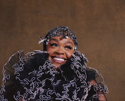 Jill Scott smiles in headshot featuring black and lavender ruffled scarf and hat/headpiece