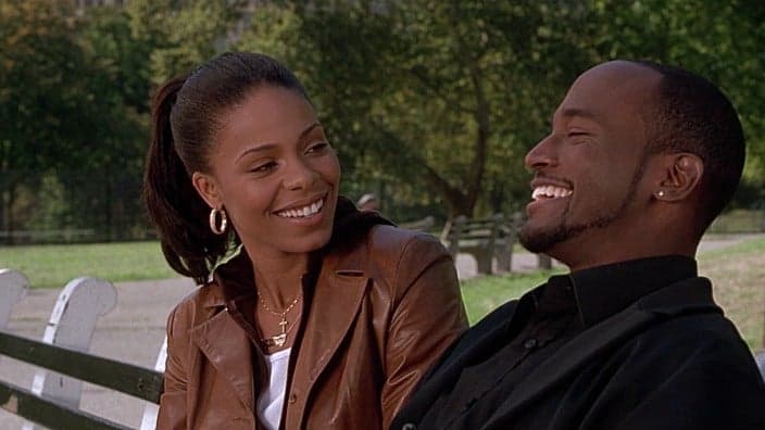 Actors Sanaa Lathan, in a brown jacket, and Taye Diggs, dressed in black, sitting on a park bench