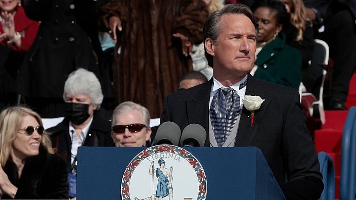 Virginia Governor Glenn Youngkin, wearing a black formal jacket, squints into the sun from behind a podium