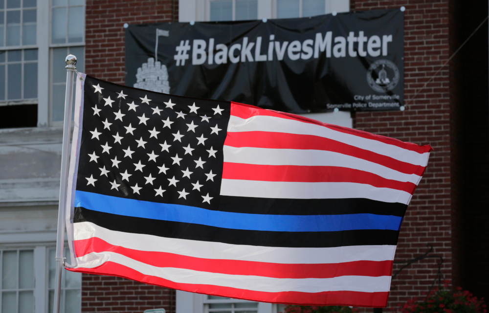 In this July 28, 2016, file photo, a flag with a blue and black stripes in support of law enforcement officers, flies at a protest by police and their supporters outside Somerville City Hall in Somerville, Mass. An Oregon county has agreed to pay $100,000 to a black employee who sued after a co-worker pinned up a "Blue Lives Matter" flag. The Oregonian/OregonLive reports Saturday, April 20, 2019, that Karimah Guion-Pledgure alleged in her January lawsuit that the flag demeans the "Black Live Matter" movement. (AP Photo/Charles Krupa, File) thegrio.com