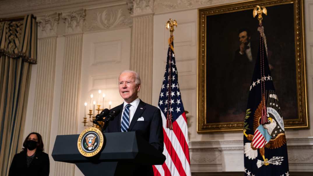 President Joe Biden stands behind a podium wearing a blue suit and blue striped tie