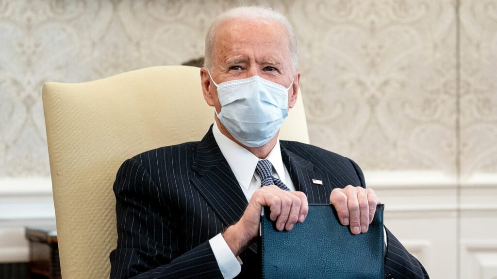 President Joe Biden sitting in a chair, wearing a medical mask and holding a blue folder