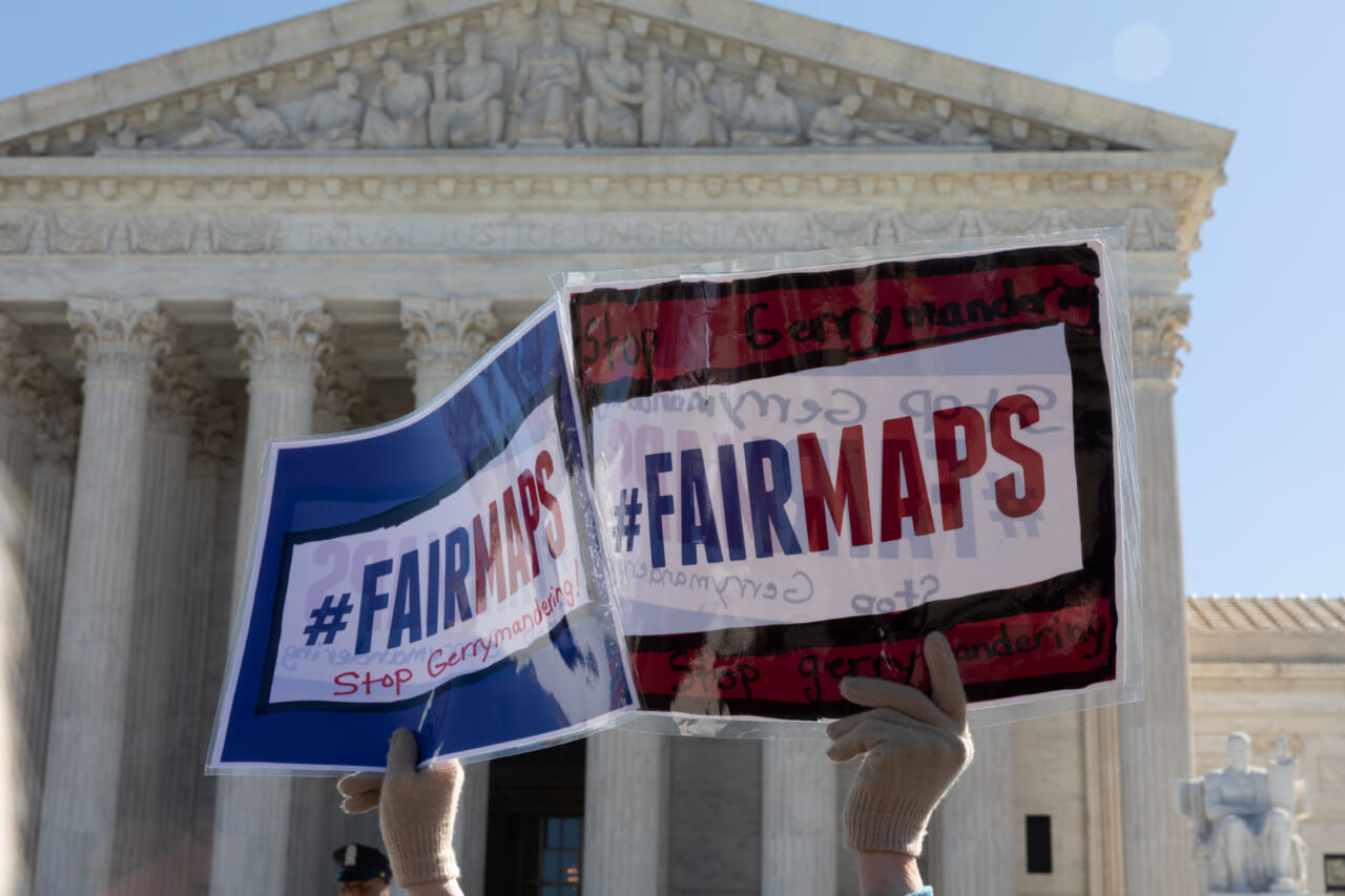 A set of hands holds up signs reading "#FairMaps - Stop Gerrymandering"