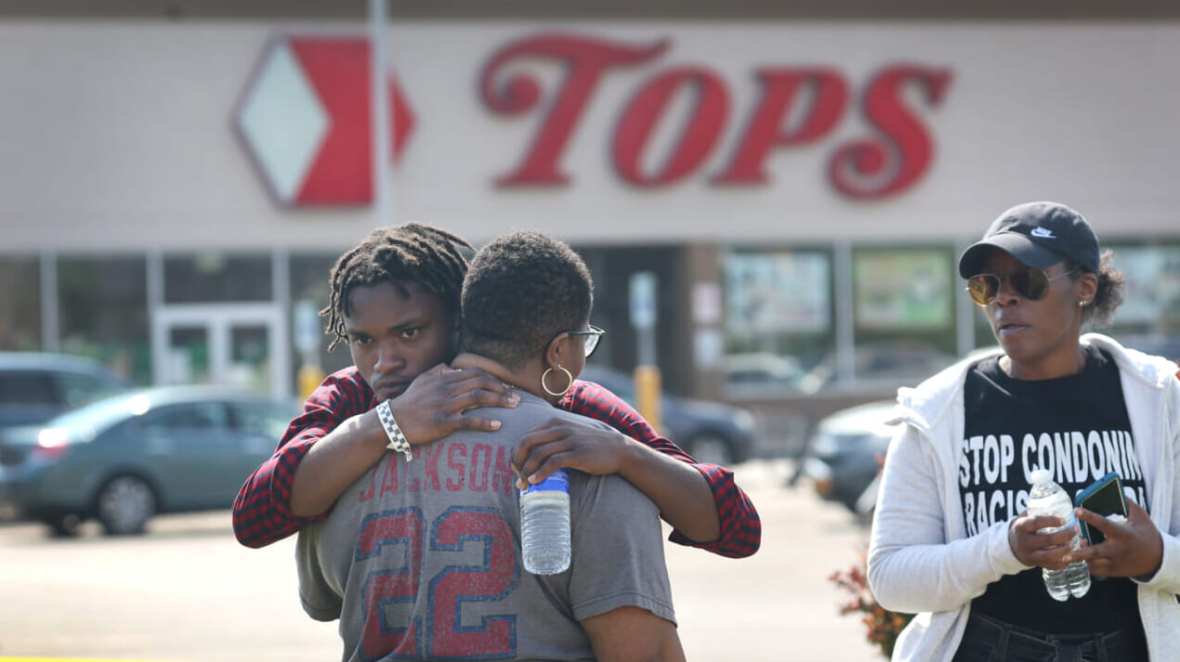 Two people embrace outside Tops market while a person holding a water bottle looks on