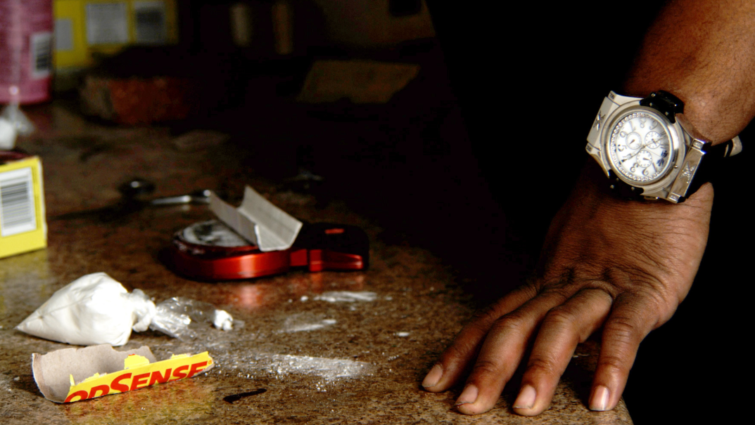 Powder, a scale and other drug paraphernalia are shown on a marble tabletop near a man's hand wearing an expensive watch