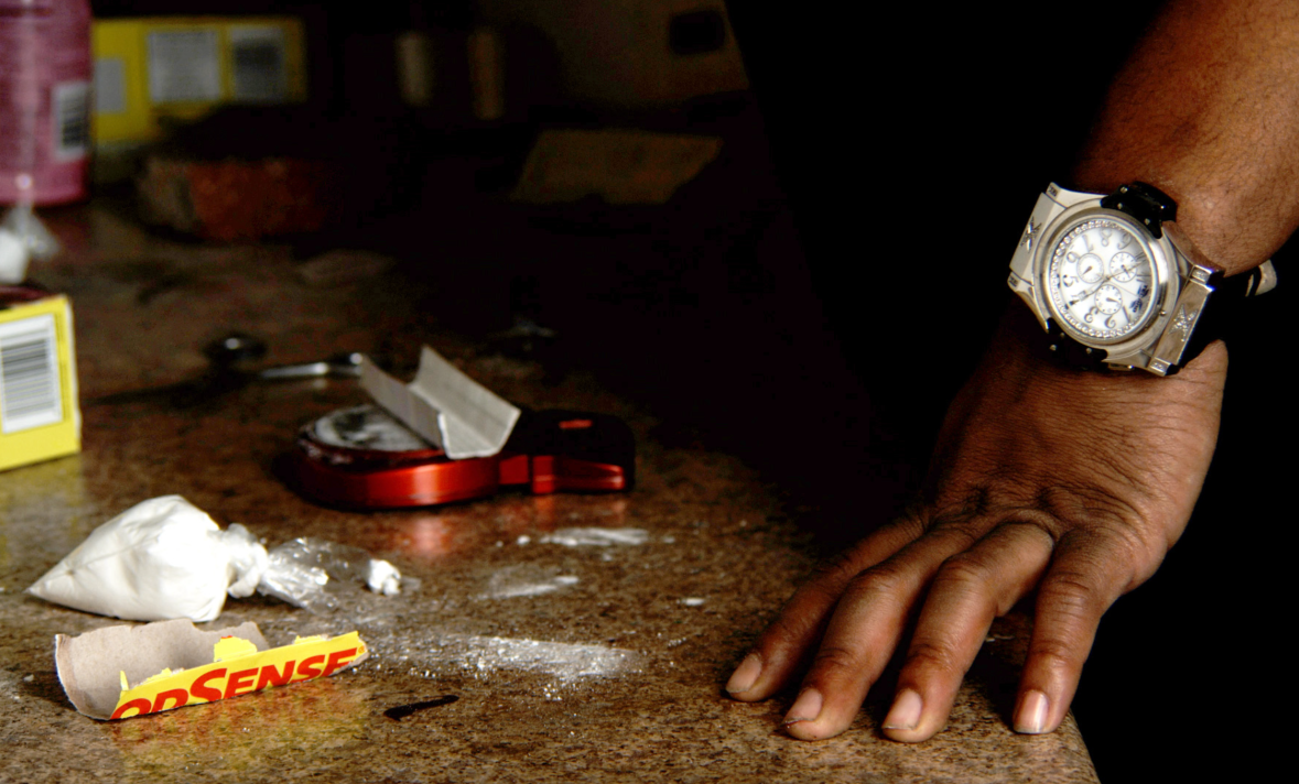 Powder, a scale and other drug paraphernalia are shown on a marble tabletop near a man's hand wearing an expensive watch