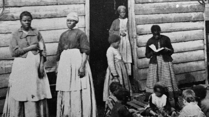 A group of women and children, presumably enslaved, sit and stand around the doorway of a rough wooden cabin, Southern United States, mid 19th Century. One girl reads a book to the group of sitting children.  Black people were enslaved by slavemasters for profit.