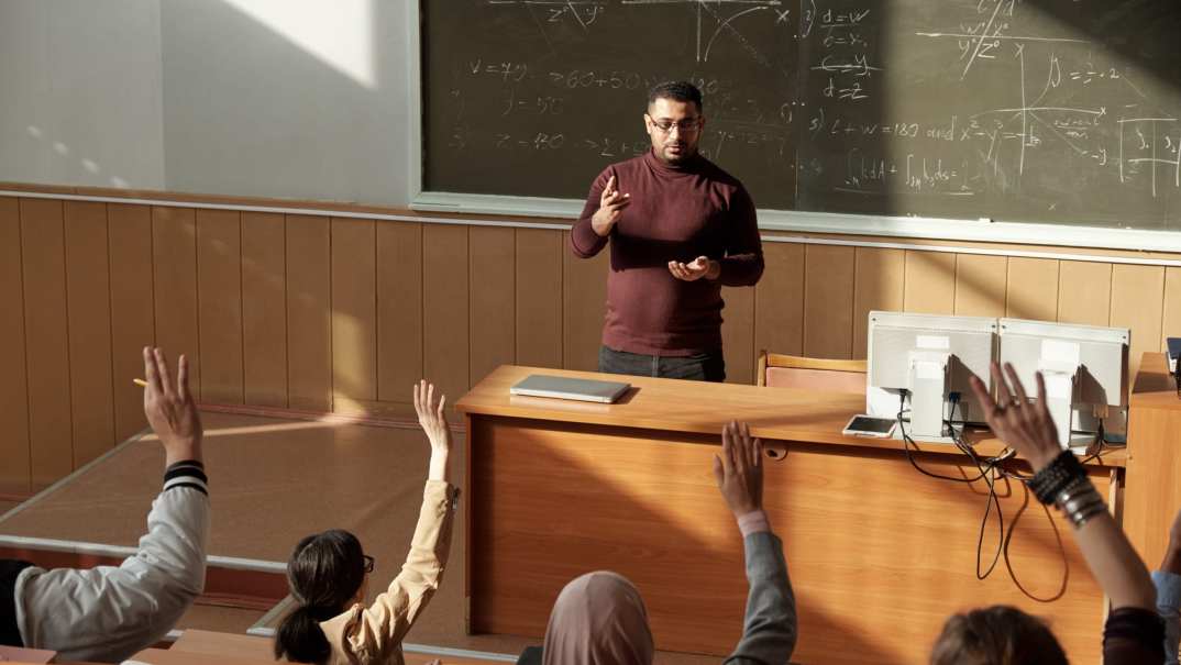 Students raise their hands as a teacher stands behind a desk