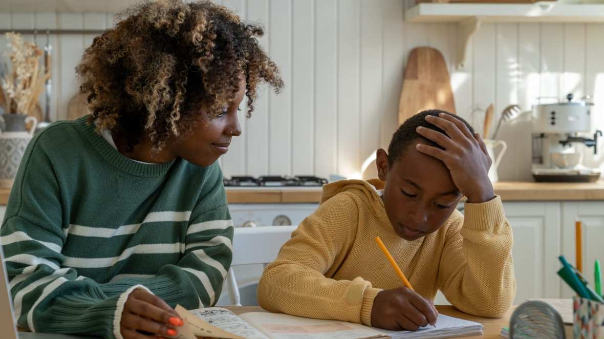 A woman watches a young boy writing in a book