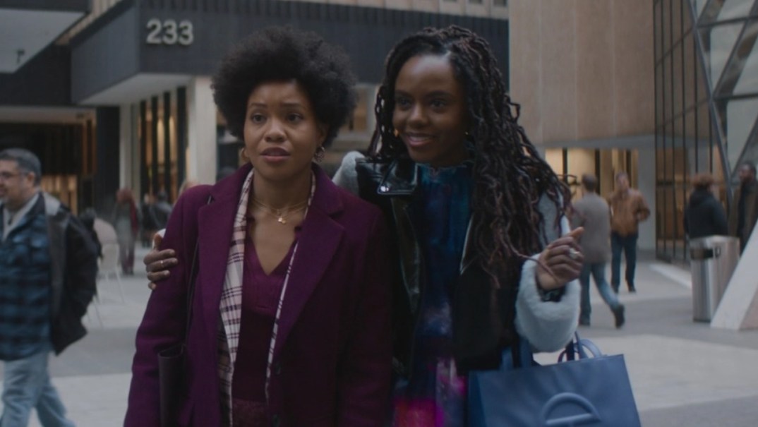 Actor Sinclair Daniel, wearing a purple coat, stands as actor Ashleigh Murray, wearing a light blue coat, holds her arm