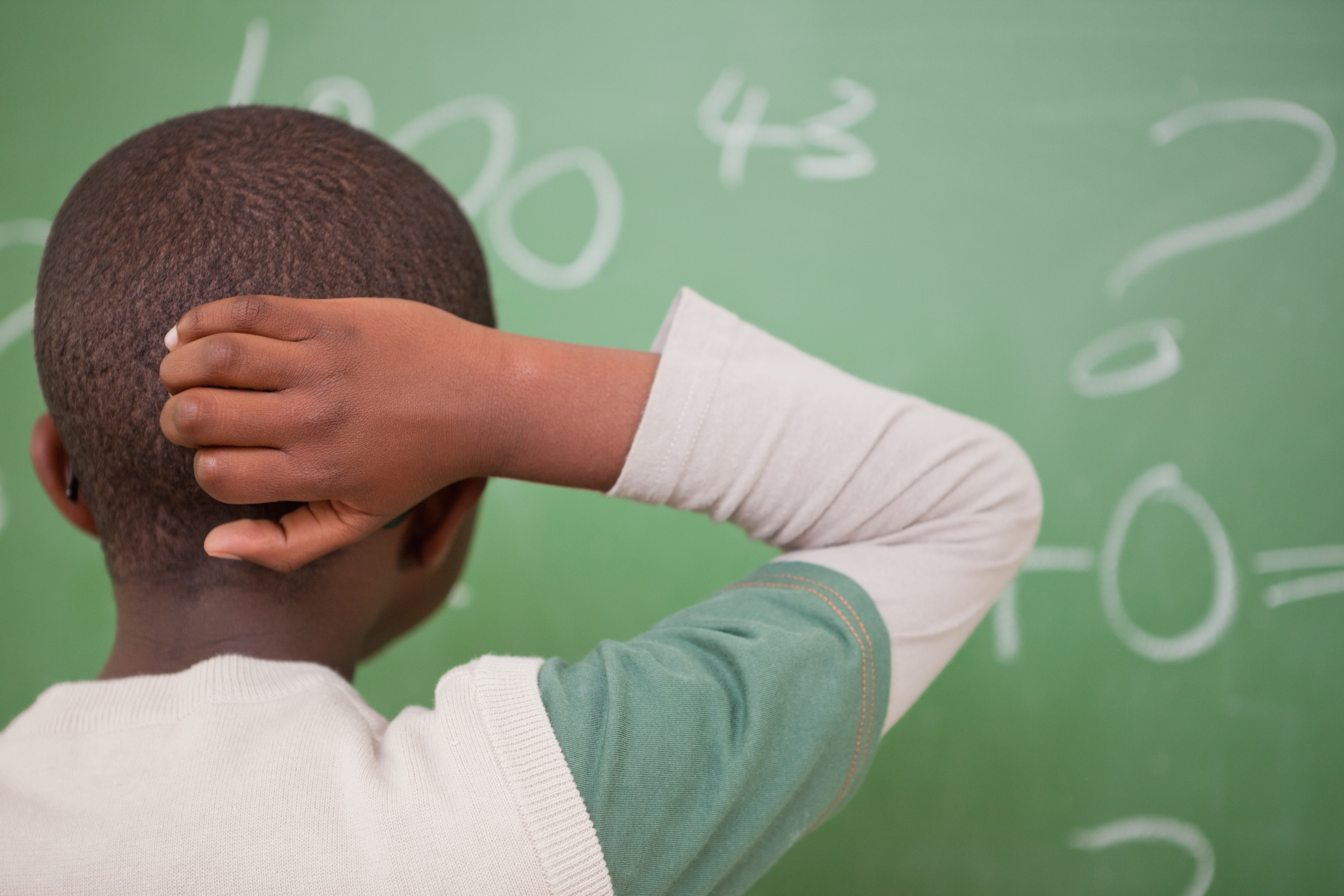 A young boy scratches the back of his head while looking at a green chalkboard