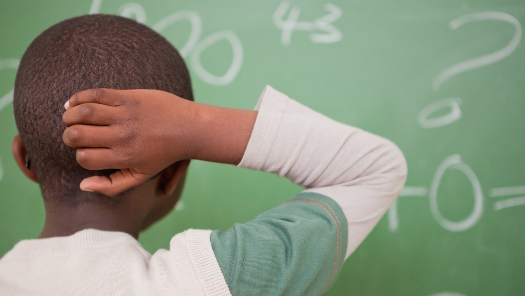 A young boy scratches the back of his head while looking at a green chalkboard