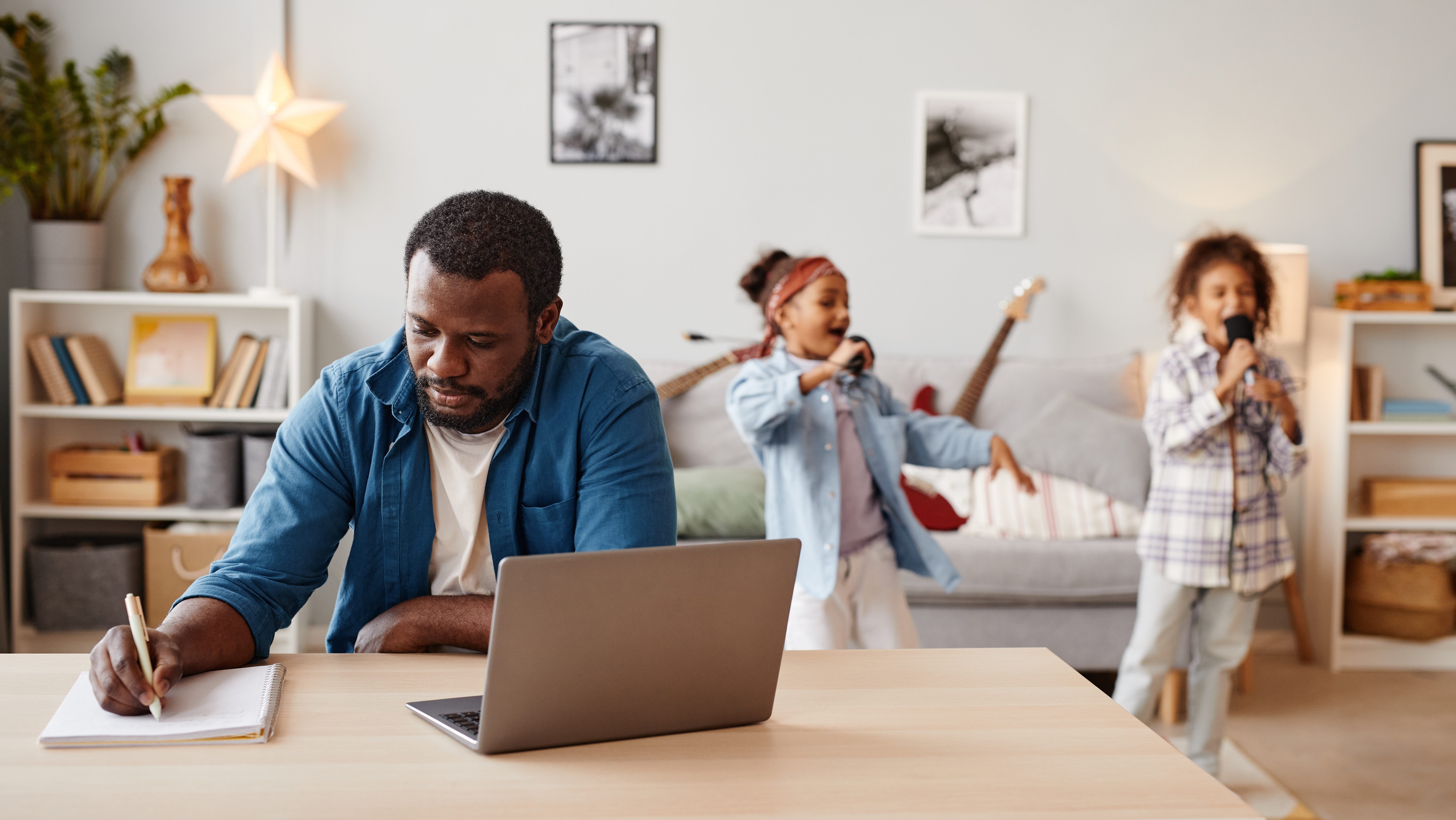 A man works at a desk with a laptop while two children play behind him