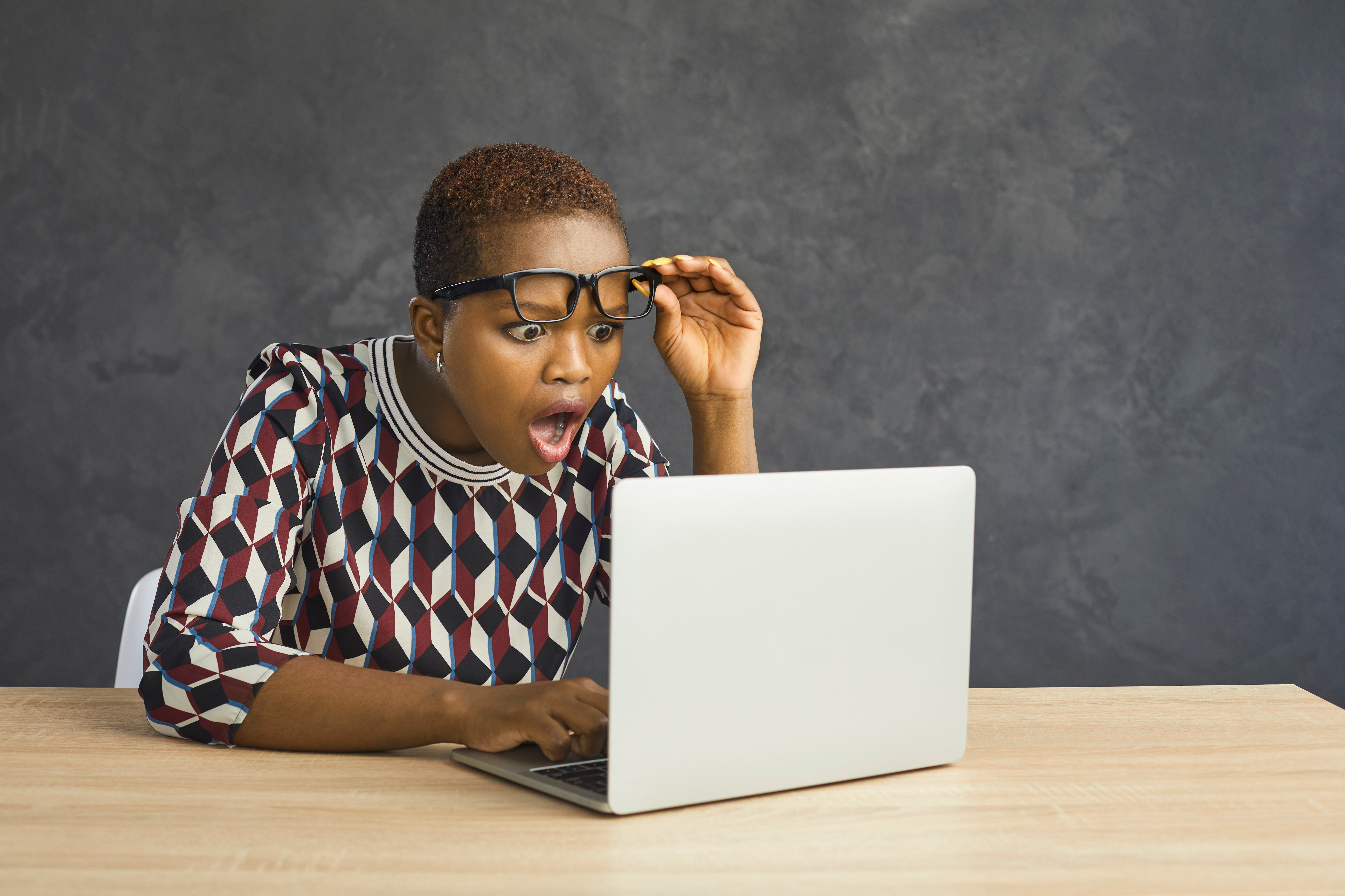 A woman lifts her glasses as she looks at a laptop screen in surprise